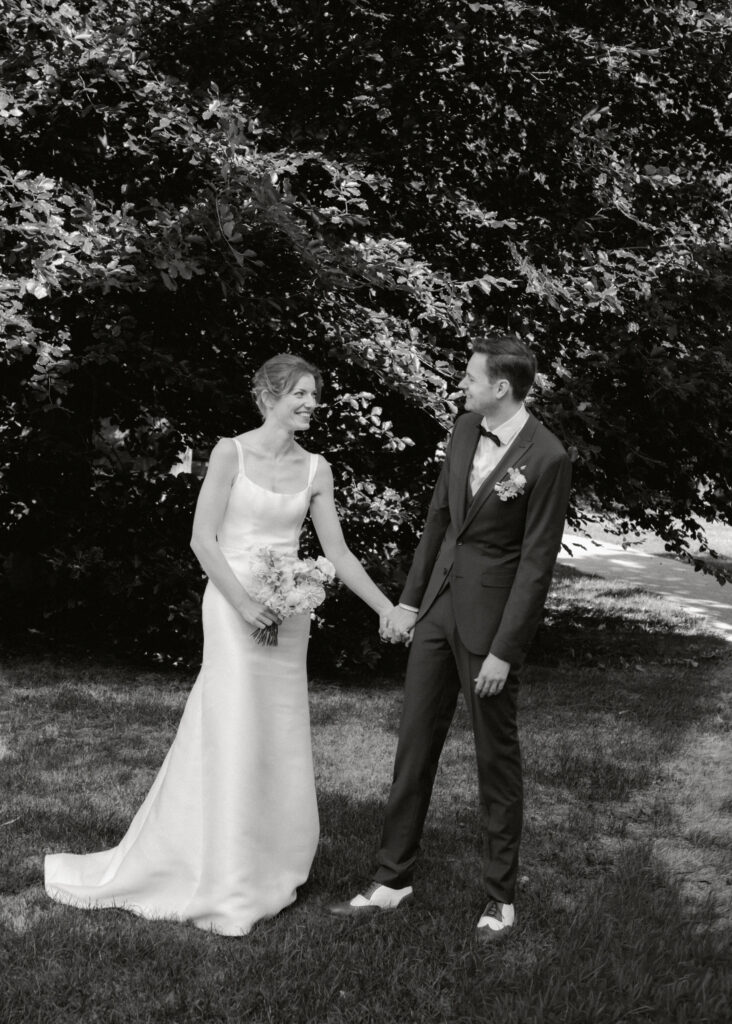 Brautpaar hält Hände und lächelt sich auf Wiese vor Bäumen an, Schwarz-Weiß-Hochzeitsfotografie von emilys stories