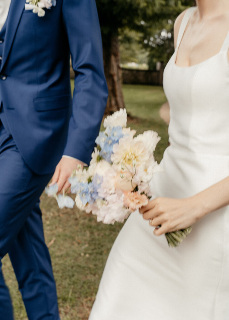 Braut im weißen Kleid hält Brautstrauß im Gehen, Bräutigam im blauen Anzug an ihrer Seite, Hochzeitsfotografie von emilys stories