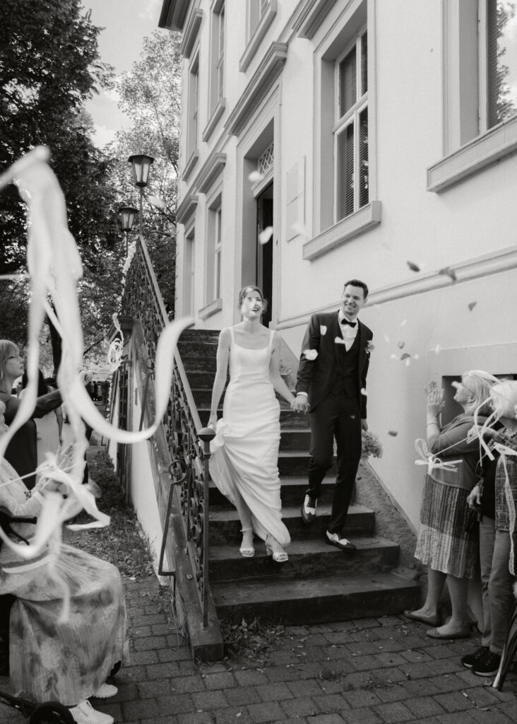 Brautpaar geht handinhand eine Treppe hinunter, Gäste werfen Konfetti, Schwarz-Weiß-Hochzeitsfotografie